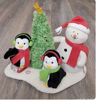 Full view of the Hallmark Jingle Pals trio on white snow-like base: green Christmas tree with star, snowman with red hat/scarf, two penguins with earmuffs and scarves