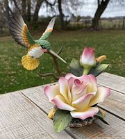 Side view of the hand-painted porcelain figurine showing the pink rose and the hummingbird in flight. Outdoor background.