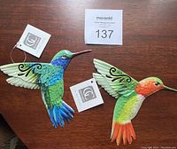 Two metal hummingbird decorations on wood table showing both with tags and detailed colors.