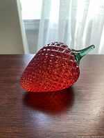 Glass strawberry paperweight sitting on a wooden surface, showing the textured red body and green stem with natural light through a window in the background.