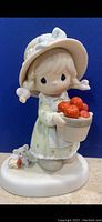 Front view of porcelain figurine featuring girl holding basket of apples and small dog at her feet against blue background.
