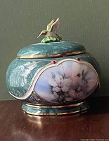 Front side view of the round porcelain music box showing 22k gold trim, Swarovski crystals, painted hummingbird and floral artwork.