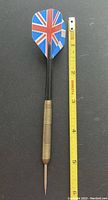 Single brass dart displayed next to measuring tape showing approx. 5 inches length. Union Jack flight design and steel point visible.