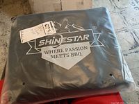 Photo showing Shinestar BBQ grill cover in unopened plastic packaging with brand logo and slogan visible