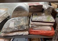 Various piled bed linens including sheets, mattress pads, and duvet covers in packaging with assorted colors including blue, white, orange, and patterns.