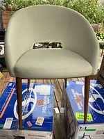 Front view of the light cream vinyl upholstered mid century modern style chair with wood legs, placed on two stacked boxes outdoors on a deck.