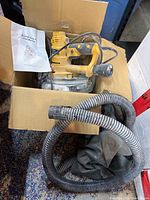 Top view of drywall sander in cardboard box with user manual and flexible dust collection hose coiled beside it.