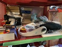 Assortment of items including white neck fans, lithium batteries, a fitness tracking sports watch, thermostats, and earbuds on metal shelves.