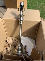 Three images showing a brushed nickel table lamp removed from its box, with no lampshade attached. The lamp socket is taped and has a pull chain. The box contains packing material.