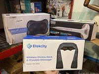 Three boxed massagers stacked on shelving showing brands and models for the lot