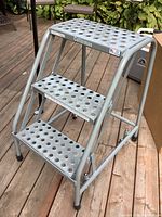 Front view of Cotterman metal step stool showing three perforated steps and sturdy frame on wooden deck.
