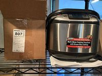 Zojirushi rice cooker placed on a black wire shelf next to a cardboard box with a label describing contents as rice cooker and Coca-Cola glasses.