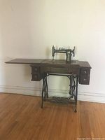 Full view of antique sewing machine mounted in wooden cabinet with side extension and drawers on iron treadle base.
