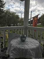 The antique ceramic tobacco jar is placed on a white quilt on a porch railing with a scenic background of trees and a flag.