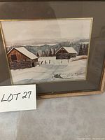 Framed art print showing two wooden barns in a snowy landscape with distant trees and hills, under glass in wood frame.