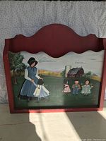 Wooden folk art organizer painted with rural farm scene, signed, showing a woman and children in a grassy field with a barn and silo in background, red curved top edge.
