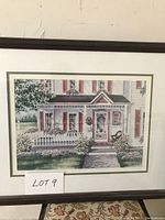 Framed print showing a colonial style house facade with porch, flowers and bushes, hung on a wall.
