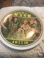 Photo of MASH 4077th commemorative plate with cast image and logo on a white background.