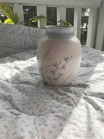Oval-shaped ceramic vase photographed on a light quilt, showing soft beige and grey colors with blue floral/leaf pattern.