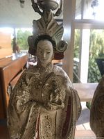 Close-up of female figurine lamp showing detailed face and ornamental object, with visible paint chipping.