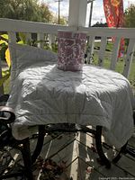Vintage metal umbrella stand shown outdoors on a porch, covered with a white cloth, with a decorative pink and white ceramic container placed on top.