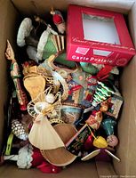 Top view of a box filled with various Christmas decorations including a straw angel ornament, ceramic Santa, wooden and pinecone ornaments, and a red Christmas postcard box.