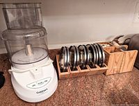 Cuisinart food processor base with clear bowl and lid beside wooden organizing boxes holding a set of stainless steel blades.