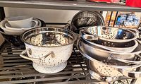 Wide view showing large metal colanders with handles, mesh strainers, and steamers stacked on a metal shelf