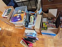 Overall view of multiple boxes and bins containing various office supplies including hole punches, tape, staplers, notebooks, and folders.