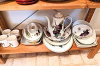 Photo showing a set of assorted kitchenware on a wooden shelf, including a teapot, covered dish, plates, saucers and cups with fruit motifs and gold trim.