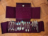 Full view of assorted silver cutlery organized in a maroon fabric roll case on a parquet floor.