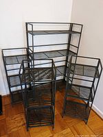 Four black metal shelving units positioned in a corner on a wooden floor. One taller shelf at back and three shorter shelves in front and sides.
