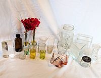 Wide view of the lot showing multiple glass shot glasses, jars, vases, a crystal block, an amber bottle, and decorative glass bowl spread on white surface