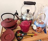 Full view of all items including cast iron red tea pot, silver kettle, red ceramic tea pot, decorative blue and red ceramic tea pot, wooden spoons, and glass pitcher