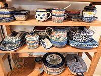 Three-tier wooden shelf holding numerous Polish pottery pieces including lidded dishes, square dishes, mugs, bowls, and a covered rectangular dish.