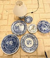 Blue and white ceramic plates and a lamp arranged on the floor showing various Spode designs including Blue Room, Castle, and Botanical patterns.