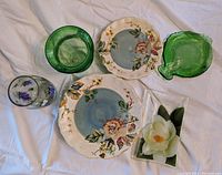 Top view of hand-painted salad and dinner plates with two green glass vases and two decorative glass trays showing floral and leaf designs.
