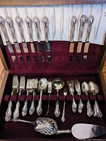 Full view of cutlery set in walnut box showing knives arranged in the lid section and various forks, spoons, and serving utensils arranged below on burgundy padded inserts.