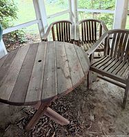 Photo showing round wooden patio table with wide plank top, central umbrella hole, and X-shaped wooden base, surrounded by three wood slatted armchairs.