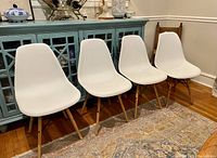 Four white molded plastic shell dining chairs with wooden legs arranged in a line against a teal cabinet, on a patterned rug.
