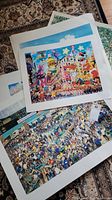 Overview of multiple art prints stacked on a carpeted floor showing colorful illustrations and city scenes.