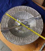 Metallic silver textured round serving platter with measuring tape showing width over 15 inches, with spoon and fork placed across the center.