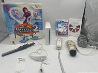Photo showing Nintendo Wii console, Dance Dance Revolution Hottest Party 2 game in box, Just Dance 3 Wii game case, and multiple accessories including sensor bar, Wii Remote with strap, Nunchuk, microphone, and cables laid out on a white surface.
