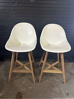 Two modern counter stools with white plastic shell seats and natural wood frames, placed side by side against a black wall.