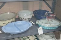 Photo showing multiple Pyrex bowls and casseroles stacked on shelf including a blue oval divided dish, mixing bowls with Butterprint and black floral patterns.