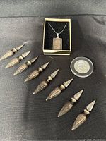 Eight sterling silver corn on the cob holders, one silver bar necklace pendant in a box, and one silver coin in protective casing on a black fabric background.