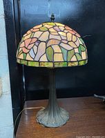 Full view of Tiffany style stained glass lamp showing floral pastel multicolor shade and bronze ornamental metal base on a wooden surface.