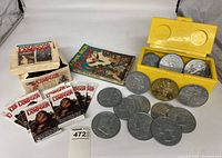 Photo showing unopened National Lampoon trading card packs in box, 1974 Comic Book Price Guide, large metal coins partly in yellow box and laid out.