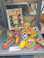 Photo showing colorful vintage tin noise makers piled next to a box of Christmas ornaments, displaying the variety of designs including a large clown face.