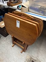 Four wooden tray tables stacked and held by a wooden stand in a vertical position, showing the warm brown wood and rounded edges.